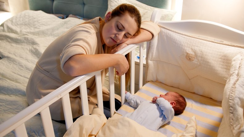 Exhausted new mother napping against baby's crib