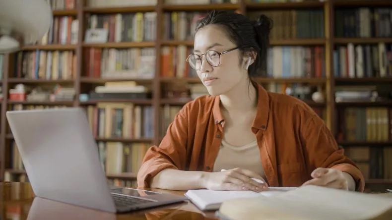 Una persona joven con gafas está sentada frente a un portátil en una biblioteca o estudio. Lleva una camisa anaranjada y tiene un cuaderno abierto, mientras parece concentrada en su trabajo o estudio. En el fondo, se ven varias estanterías llenas de libros.