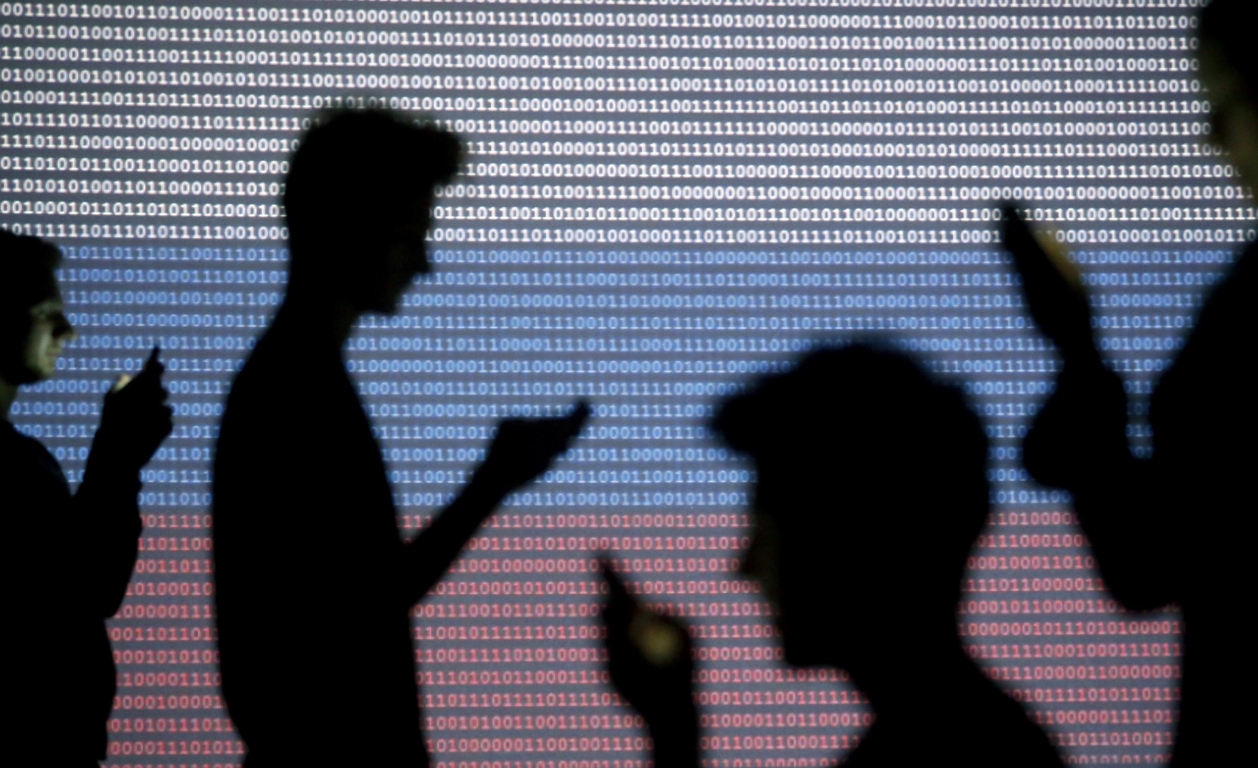 People are silhouetted as they pose with mobile devices in front of a screen projected with a binary code and a Russian national flag.