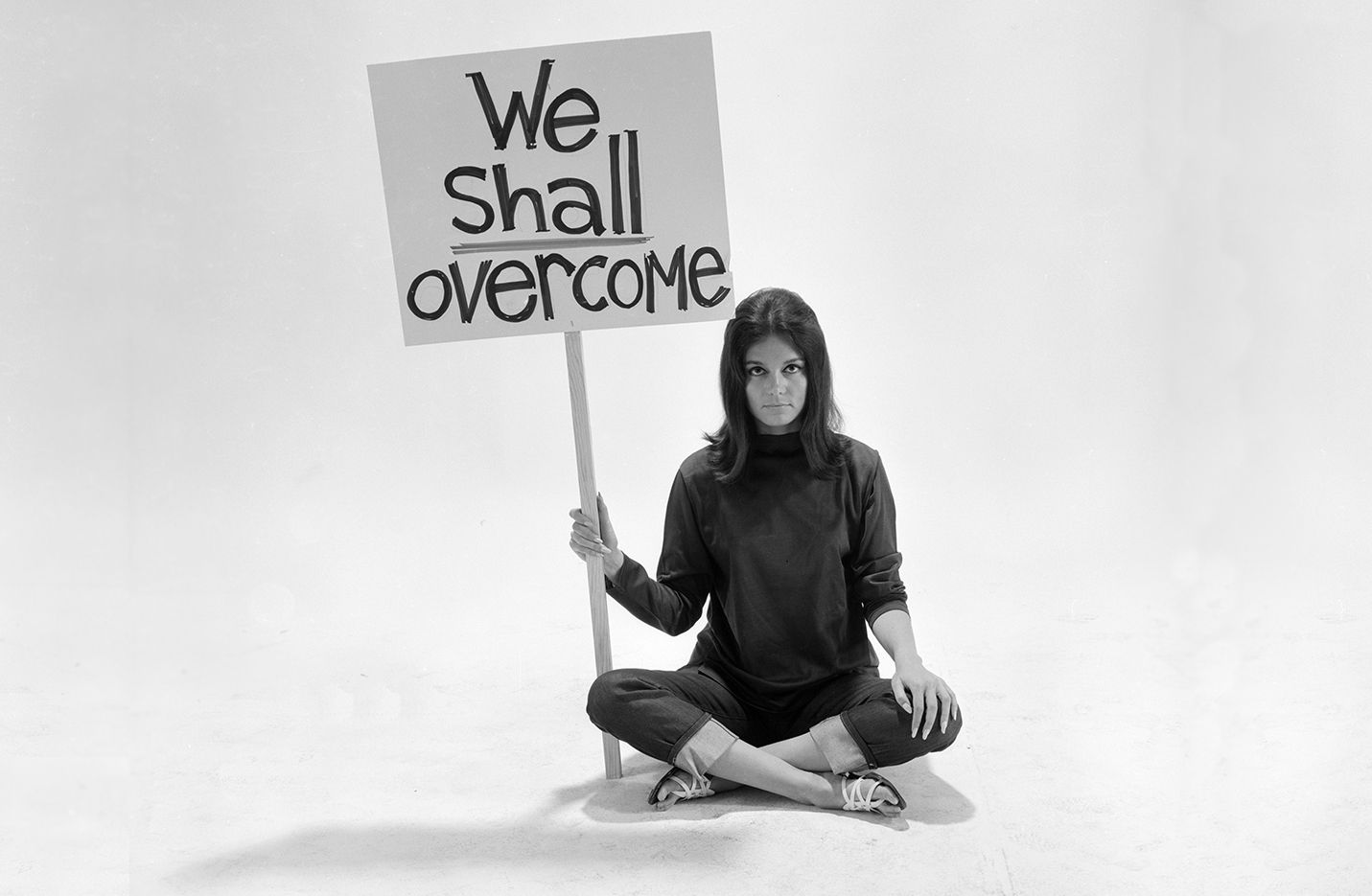photographie de Gloria Steinem en 1965, assise en tailleur, l'air sérieux et déterminé, regardant l'objectif, tenant une pancarte "We Shall Overcome" ("Nous Surmonterons")